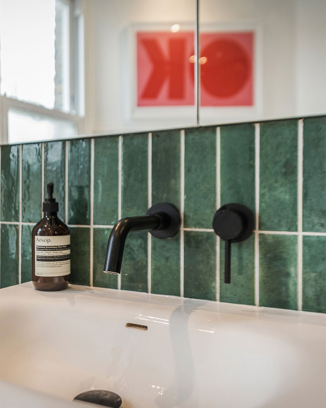Bottle of Aesop product on a bathroom sink with green tiles and black fixtures.