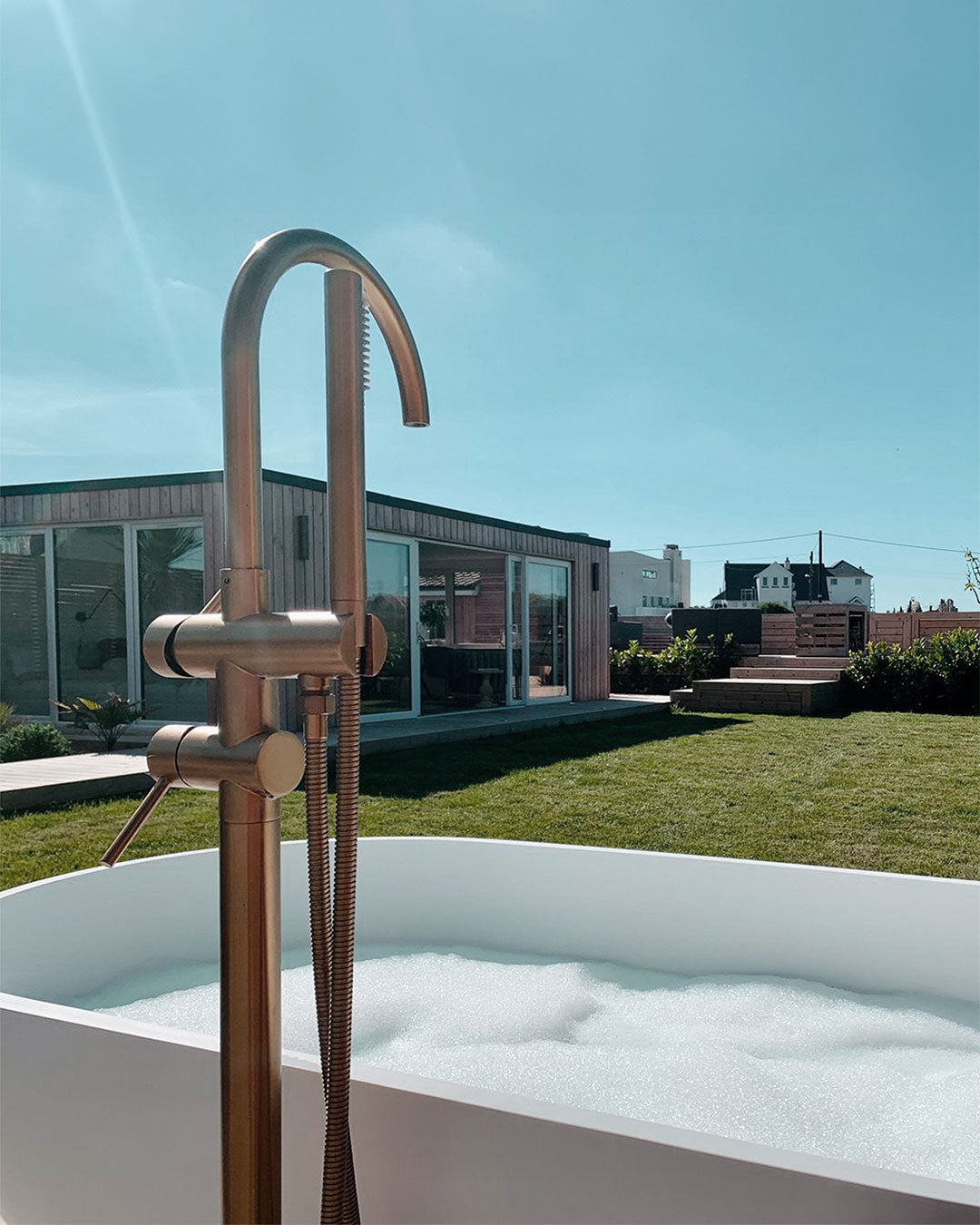 Outdoor bathtub with a bronze faucet and shower head against a clear blue sky.