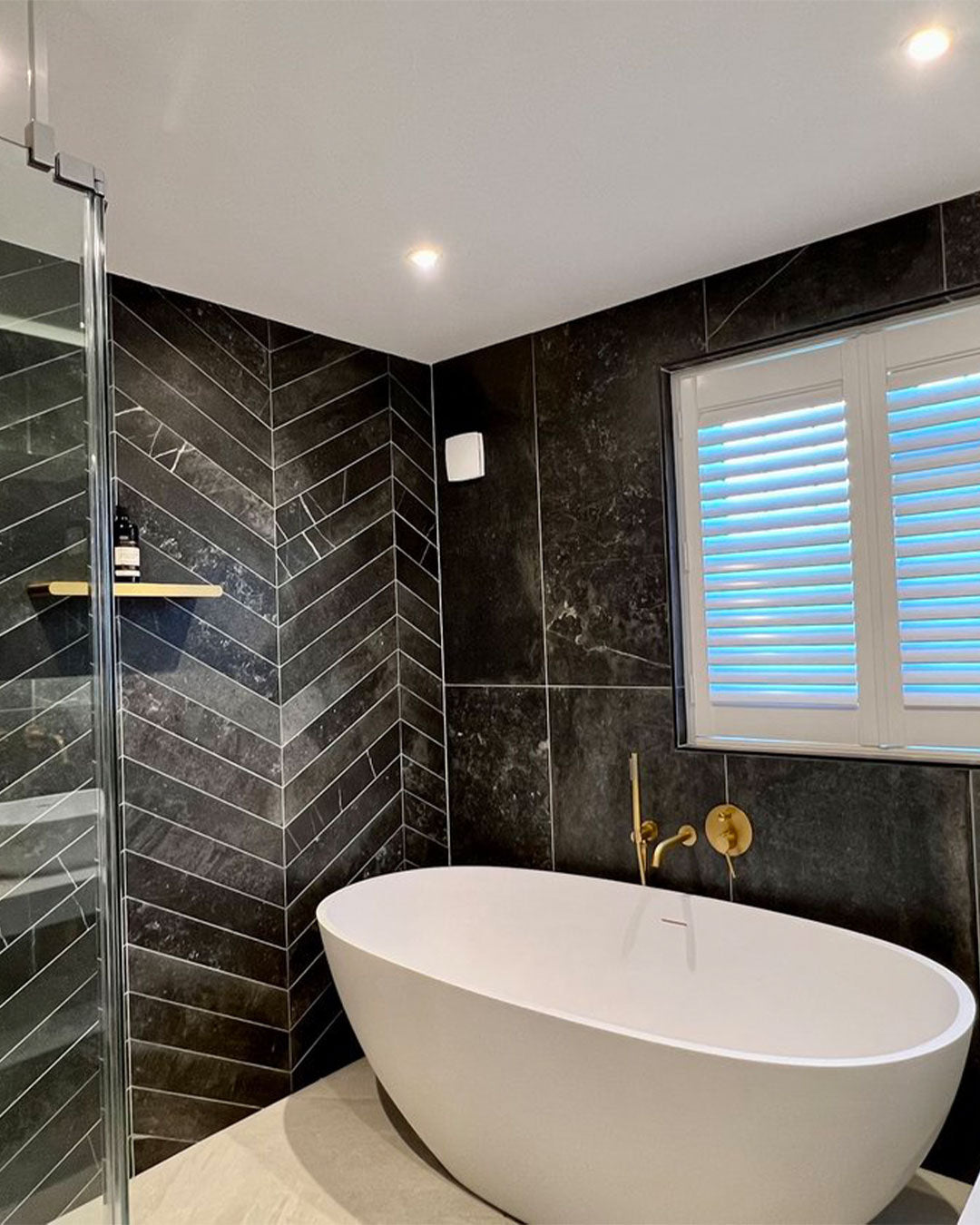 Modern bathroom with black tiled walls, a white freestanding bathtub, and a window with blue blinds.
