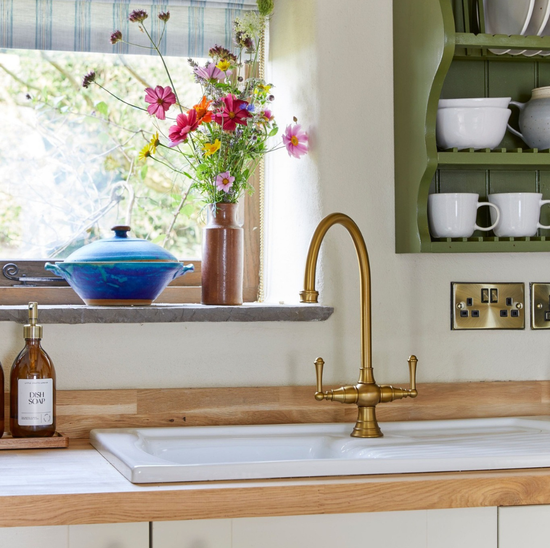 Kitchen sink with gold faucet, window with flowers, and shelves with mugs.