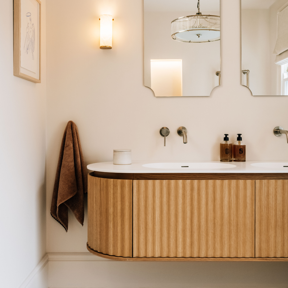 A modern bathroom vanity with a curved light wood fluted cabinet, a white sink basin, and brass fixtures, shown in a styled interior with mirrors, a wall sconce, and a hanging brown towel.