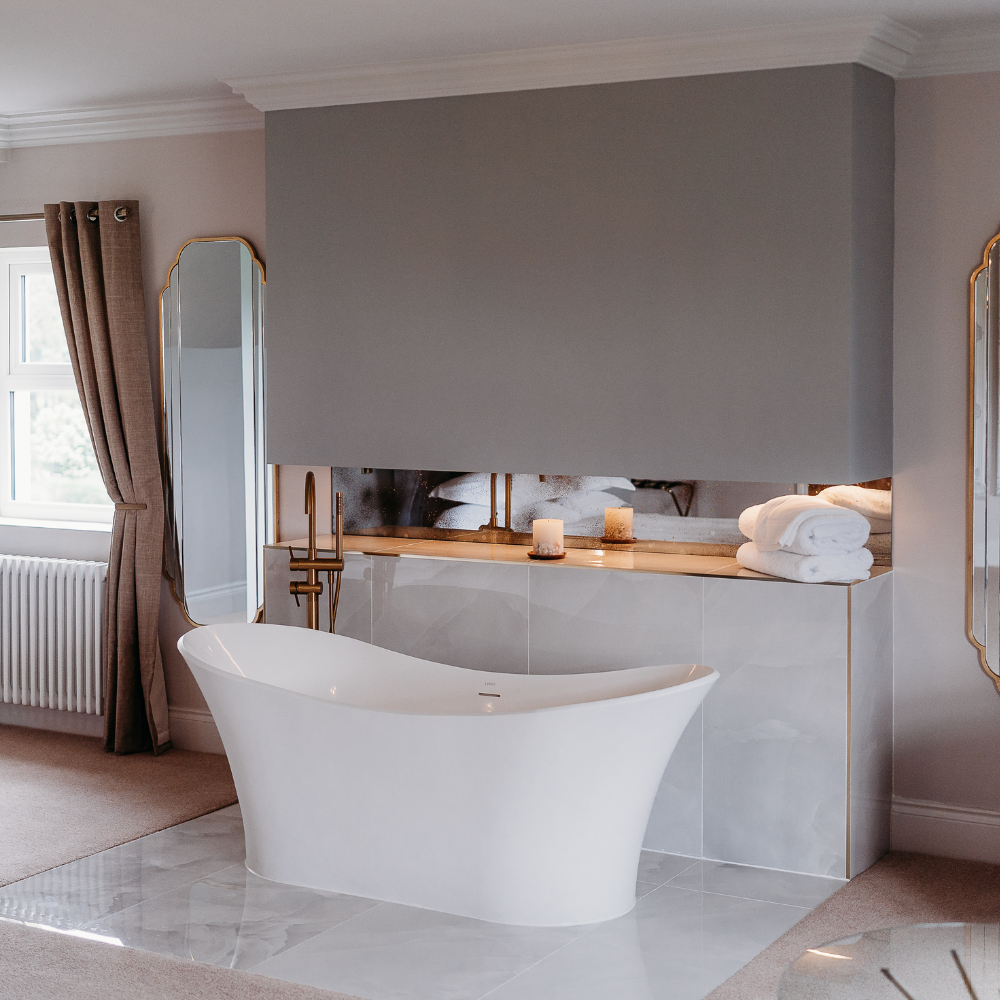 A modern freestanding white bathtub placed in a luxurious bathroom setting with a marble-tiled counter, gold-framed mirrors, and neutral beige walls.