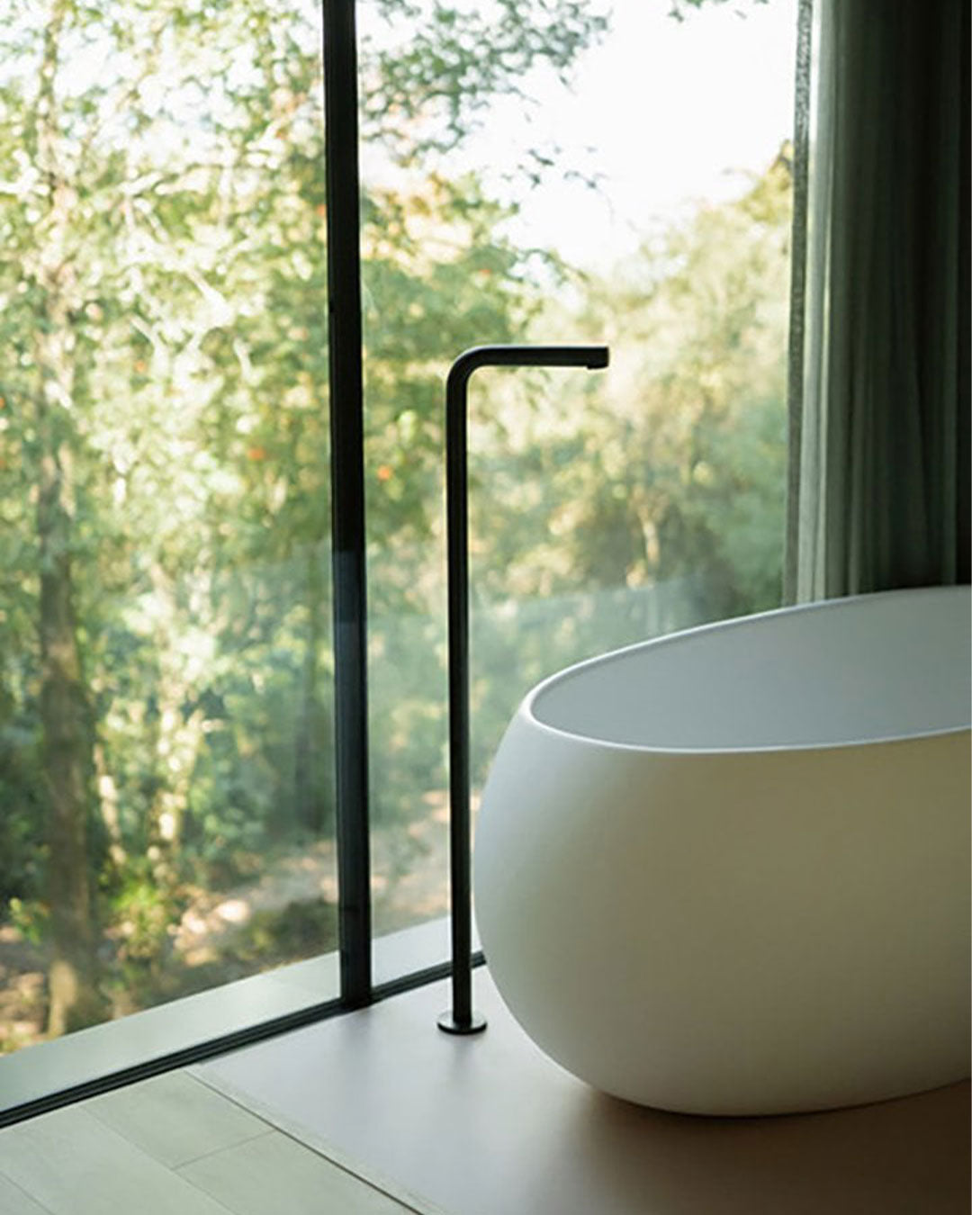 Modern white freestanding bathtub with black faucet in a bathroom with large windows showing greenery outside.