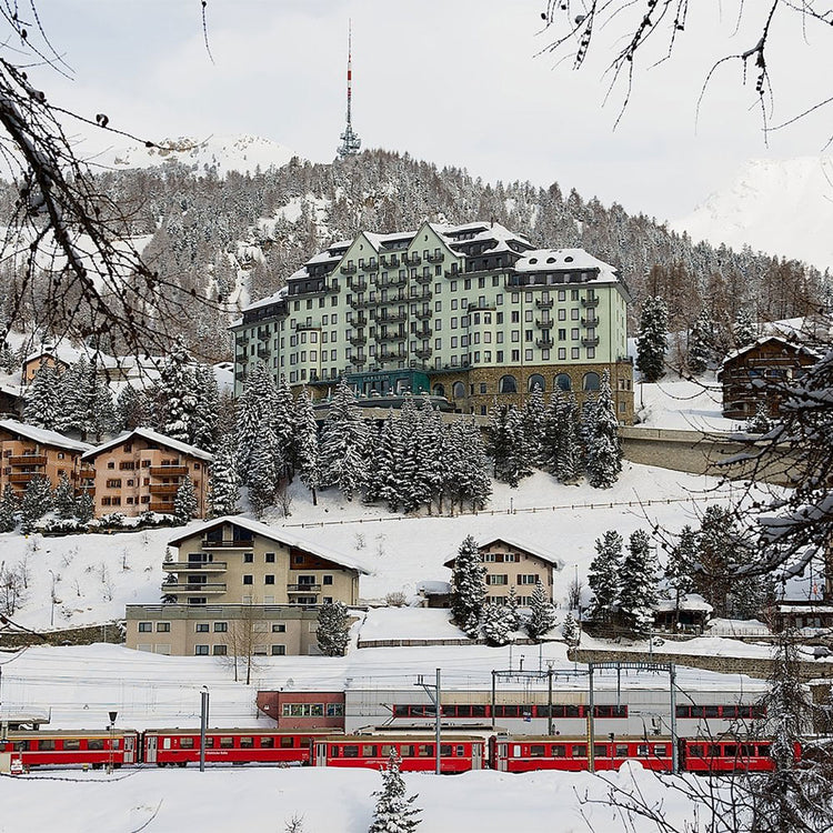Snow-covered mountain village with a large building and a red train passing by.