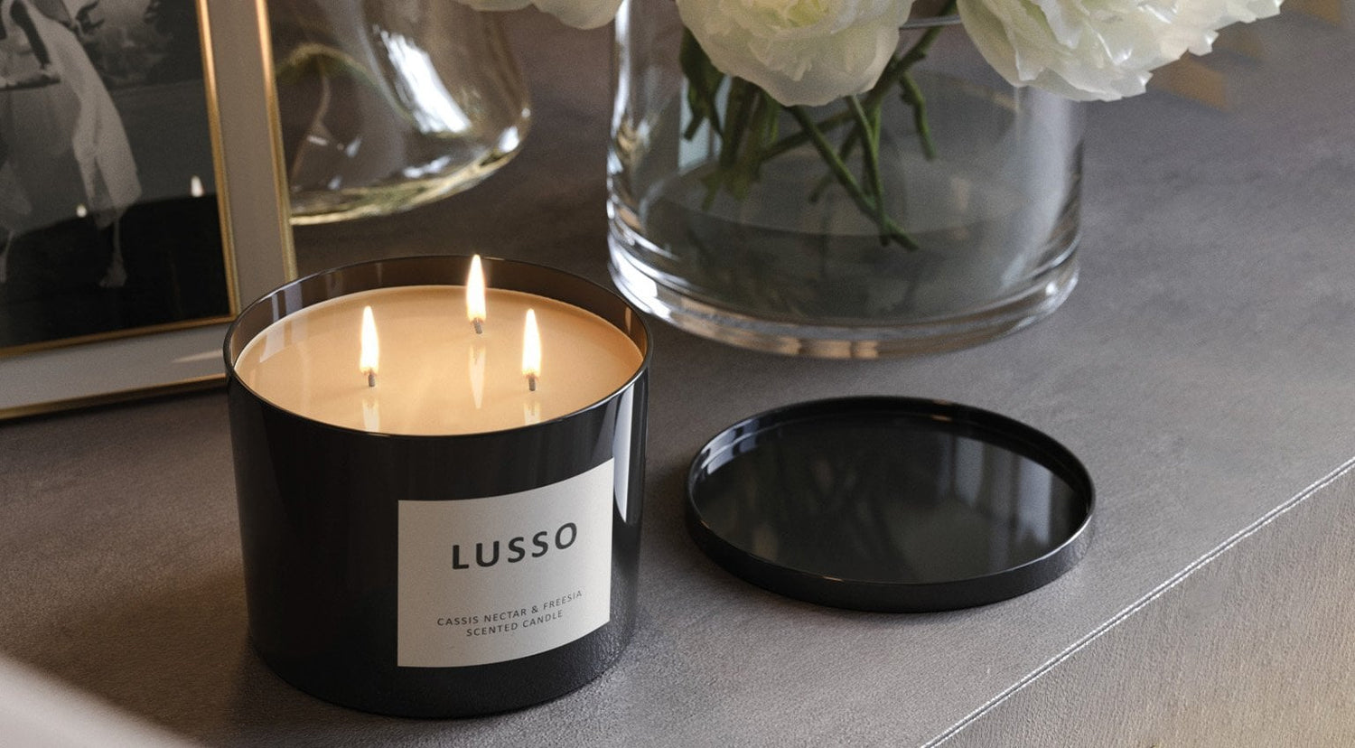 Black Lusso candle with three lit wicks on a gray surface, next to a vase with white flowers.