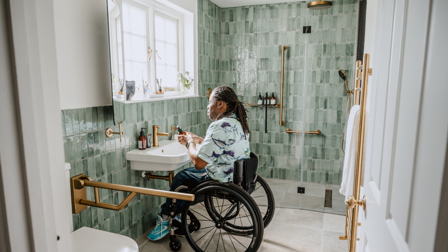 Person in a wheelchair using a sink in a bathroom with green tiled walls.