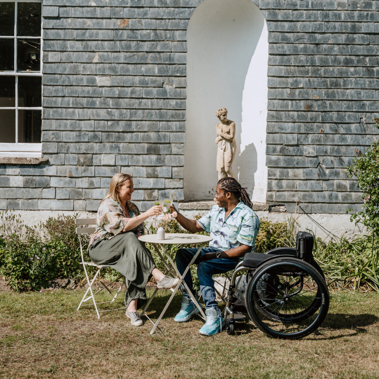 Two people sitting at a table outdoors, one in a wheelchair, with a stone building in the background.