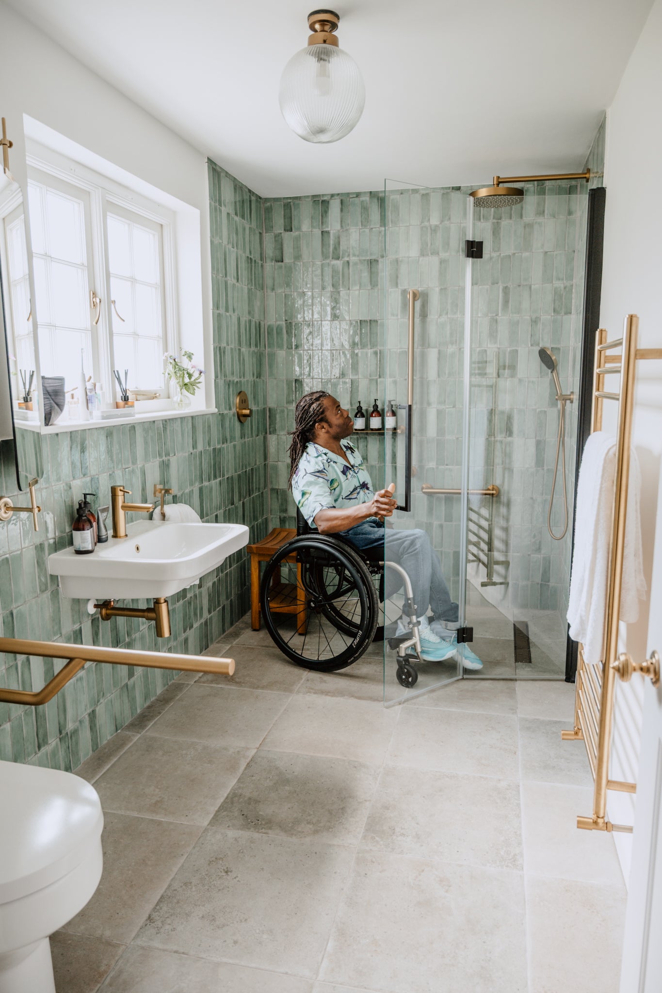 Person using a wheelchair in a modern bathroom with green tiled walls and gold fixtures.