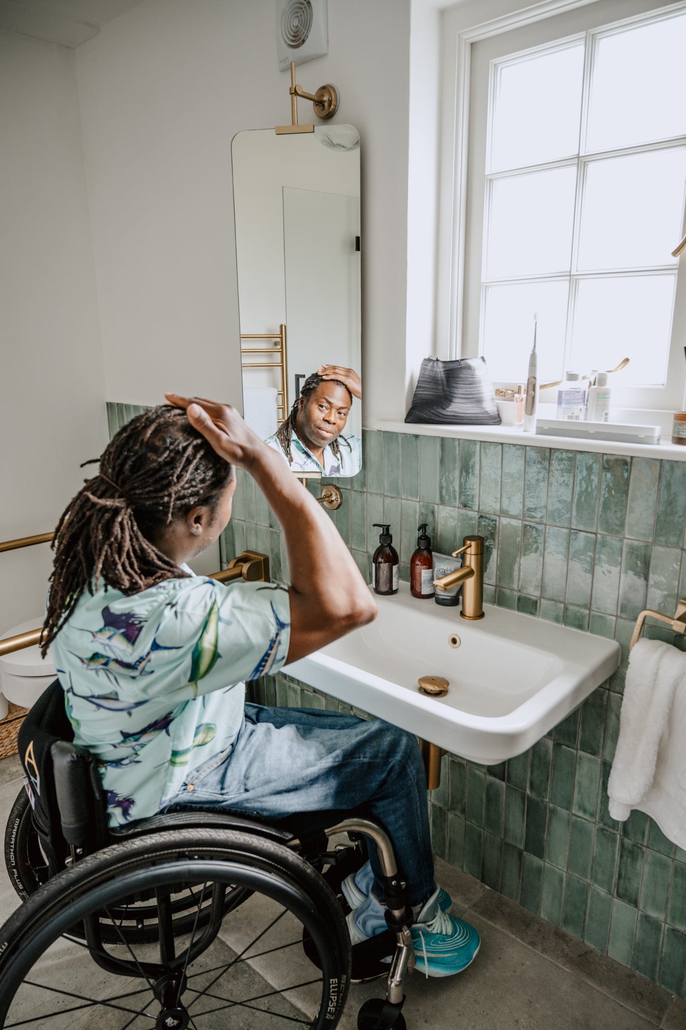 Person in a wheelchair grooming themselves in a bathroom with green tiles.